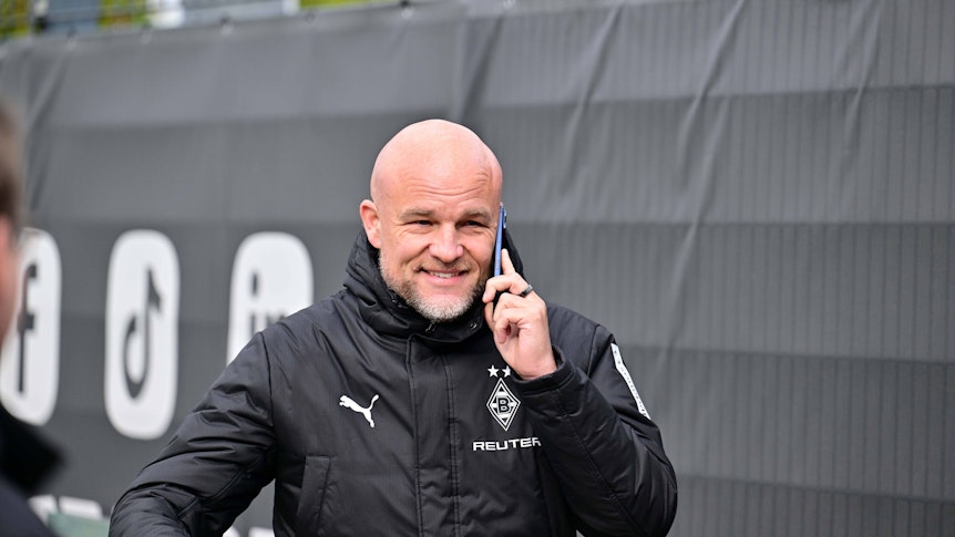Rouven Schröder telefoniert in Gladbach-Jacke am Trainingsplatz und grinst dabei.
